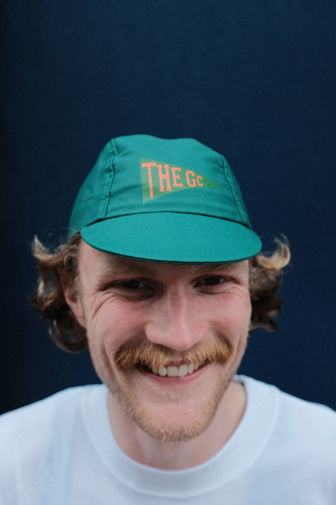 GC Flags cycling cap in green worn in front of a blue wall